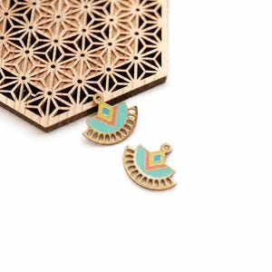 Peut inclure: Deux pendentifs dorés en forme d'éventail avec des détails en émail turquoise, corail et jaune. Un motif géométrique en bois découpé au laser est en arrière-plan. Les pendentifs ont de petites boucles.