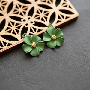 Può includere: Due ciondoli a forma di fiore verde con centri color oro e piccoli anelli per appenderli. I ciondoli sono appoggiati su una superficie grigio scuro accanto a un elemento decorativo in legno con un motivo geometrico.