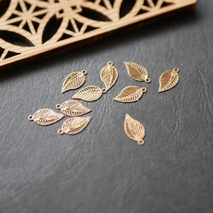 Puede incluir: Una colección de pequeños dijes dorados con forma de hoja. Cada dije presenta vetas detalladas y un pequeño lazo para sujetar. Los dijes están dispersos sobre una superficie gris oscuro, con un elemento decorativo de madera en el fondo.