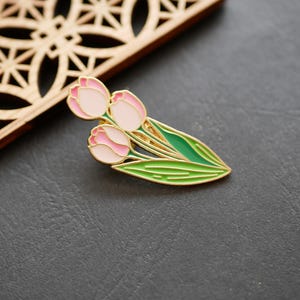 以下が含まれることがあります： 金色の金属製ピン。ピンクと白のチューリップの花が3つ、緑の葉が付いています。ピンは金色の縁取りが施された詳細なデザインで、濃い灰色の背景に配置されています。