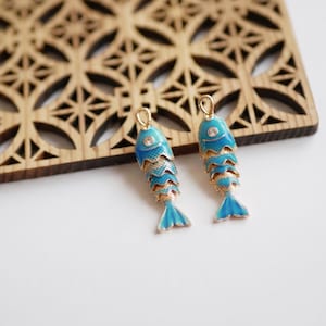 Può includere: Due ciondoli a forma di pesce blu e oro con uno sfondo in legno decorativo. I ciondoli hanno un design a scaglie dettagliato e una piccola gemma sulla testa. I ciondoli a forma di pesce sono di un blu vibrante con accenti dorati.