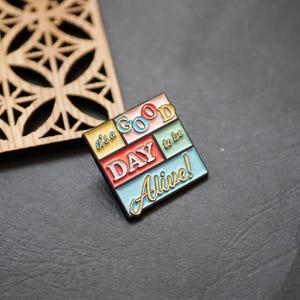 May include: A colourful enamel pin with the text "It's a good day to be alive!" in a retro style. The pin is made of metal and has a black backing.