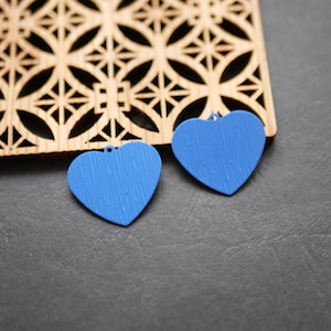 Peut inclure: Deux pendentifs bleus en forme de cœur avec une surface texturée. Les pendentifs ont un petit trou en haut pour être suspendus. Une pièce décorative en bois avec un motif géométrique est en arrière-plan. Les pendentifs sont sur une surface gris foncé.