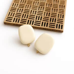 May include: Two cream-colored, smooth, rectangular jewelry components with rounded corners. Each piece features a small, gold-toned metal loop at the bottom edge, designed for attaching additional elements to these flat, solid-colored items.