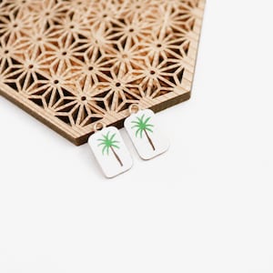 Puede incluir: Dos pendientes rectangulares blancos pequeños con un diseño de palmera verde y aros dorados. Los pendientes están junto a una pieza decorativa de madera con un patrón geométrico. El fondo es blanco.
