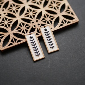 Könnte beinhalten: Zwei rechteckige Ohrringe mit weißem Hintergrund und einem schwarzen Blattdesign. Die Ohrringe haben einen goldenen Rand und einen kleinen schwarzen Punkt oben. Sie werden auf einem dekorativen Holzstück mit geometrischem Muster präsentiert.