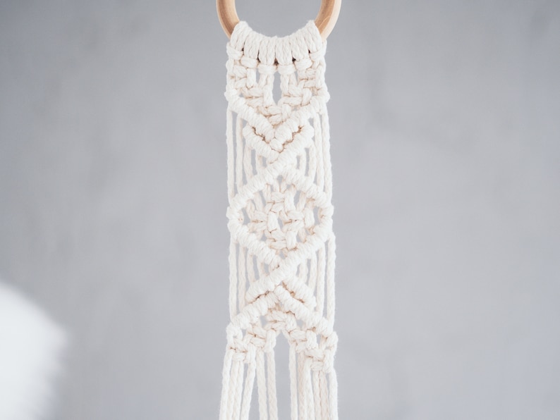 Puede incluir: Un macetero de macram&eacute; blanco con un anillo de madera en la parte superior. El macetero est&aacute; hecho con una t&eacute;cnica de anudado que crea un patr&oacute;n geom&eacute;trico.