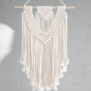 May include: A white macrame wall hanging with a geometric design. The hanging is made of cotton cord and has a wooden dowel at the top.