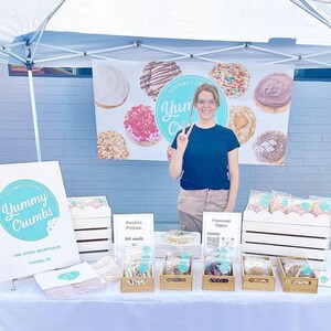 Puede incluir: Una mujer se encuentra detr&aacute;s de una mesa con un cartel que dice "Gourmet Cookies Yummy Crumbs and other Delectables Tucson, AZ". La mesa est&aacute; preparada con varias galletas en bolsas de pl&aacute;stico transparentes, dispuestas en cajas de madera. Las galletas est&aacute;n decoradas con coloridos espolvoreados y glaseado. Un cartel detr&aacute;s de la mesa dice "Gourmet Cookies Yum Crumbs".