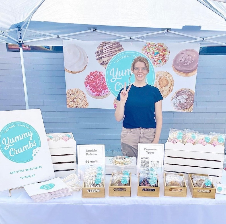 Puede incluir: Una mujer se encuentra detr&aacute;s de una mesa con un cartel que dice "Gourmet Cookies Yummy Crumbs and other Delectables Tucson, AZ". La mesa est&aacute; preparada con varias galletas en bolsas de pl&aacute;stico transparentes, dispuestas en cajas de madera. Las galletas est&aacute;n decoradas con coloridos espolvoreados y glaseado. Un cartel detr&aacute;s de la mesa dice "Gourmet Cookies Yum Crumbs".