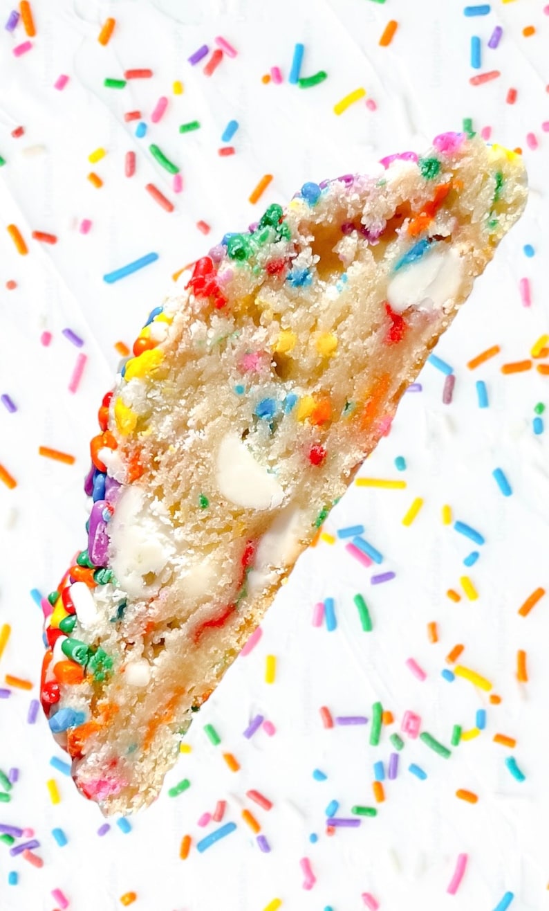Puede incluir: Primer plano de una galleta a medio comer con chispas de chocolate blanco y chispas de arco&iacute;ris. La galleta est&aacute; sobre un fondo blanco con m&aacute;s chispas de arco&iacute;ris.