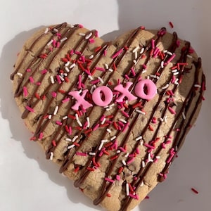 May include: A heart-shaped chocolate chip cookie decorated with chocolate drizzle, pink and red sprinkles, and pink fondant letters spelling "XOXO".