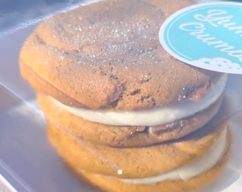 May include: A double-decker cookie sandwich with a light brown cookie and white frosting. The cookie is topped with sugar crystals. The cookie is in a clear plastic container with a blue and white sticker that says "Yum Crumbs".