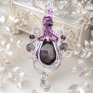 Peut inclure: Un pendentif en fil métallique torsadé avec une pierre ovale violet foncé au centre, entourée de fils argentés et violet vif. Le design intègre de petites perles violettes à facettes et des motifs en spirale complexes et symétriques.