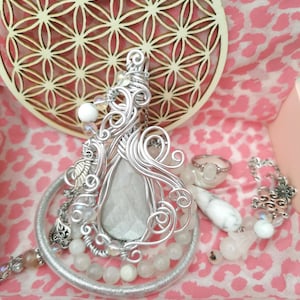 Op de afbeelding: Een verzameling handgemaakte sieraden, waaronder een grote zilveren hanger met een druppelvormige steen, een ring en een kralenarmband. De hanger is ingewikkeld met draad gewikkeld. De achtergrond heeft een roze luipaardprint.