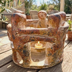 May include: A circular terracotta candle holder featuring a ring of stylized figures with arms linked around the perimeter. The clay has a textured, mottled finish in shades of brown, ochre, and black, with a small lit candle placed in the center.