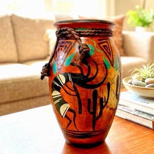 May include: A decorative ceramic vase featuring a warm orange and brown base color with hand-painted black Kokopelli figures, cactus motifs, and geometric patterns. The neck of the vase is wrapped with a thick, dark brown twisted rope accent.