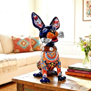 May include: A ceramic dog figurine featuring a dark blue head, large ears with red and blue floral patterns, and a body decorated with vibrant orange, blue, and yellow geometric designs. The dog has a brown collar and holds a white bone in its mouth.