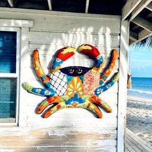 May include: A colorful crab wall decoration with a vibrant design. The crab features red, yellow, blue, and orange accents. It is mounted on a white wooden wall, with a beach and ocean visible in the background.