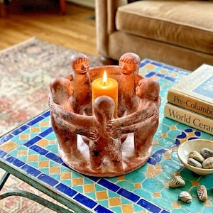 May include: A circular terracotta candle holder featuring four stylized figures standing in a ring with their arms linked. The figures are reddish-brown in color and surround a single yellow pillar candle placed in the center of the sculpture.
