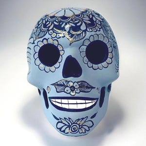 May include: A light blue decorative skull with intricate dark blue and gold floral designs. The skull features large eye sockets, a dark blue nose, and a smiling mouth with white teeth. The design is symmetrical and detailed.