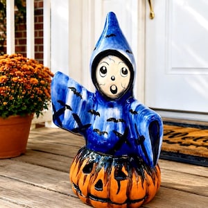 May include: A ceramic ghost figurine with a blue hooded cloak featuring black bat silhouettes, emerging from a carved orange pumpkin base. The ghost has a white face with large black eyes and an open mouth expression, standing on a wooden surface.