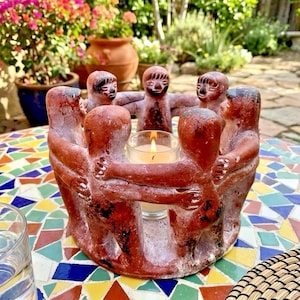May include: A circular terracotta candle holder featuring a ring of six stylized figures with arms linked together. The reddish-brown clay figures surround a central glass votive cup containing a lit white candle with a visible flame.