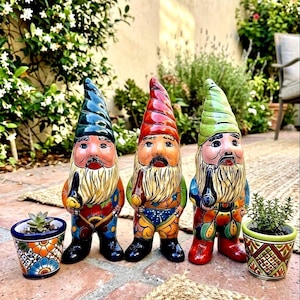 May include: Three ceramic garden gnomes with pointed hats in dark blue, red, and light green, each featuring a long beard and a pipe. They are accompanied by two small, colorful hand-painted ceramic pots, each containing a small green succulent plant.
