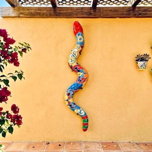 May include: A long, serpentine wall sculpture featuring a vibrant, multi-colored Talavera-style pattern with red, blue, yellow, and green hues. The snake-shaped piece is mounted vertically, displaying a glossy finish and intricate floral motifs.