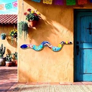 May include: A decorative ceramic snake wall sculpture featuring a vibrant, multi-colored floral pattern in shades of blue, yellow, red, and green. The elongated, undulating form is mounted horizontally on a textured, warm-toned stucco wall surface.