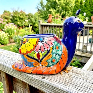May include: A ceramic snail-shaped planter featuring a glossy dark blue head and neck. The shell is decorated with vibrant, hand-painted floral patterns in shades of yellow, pink, green, and orange, with a wide opening at the top for plants.