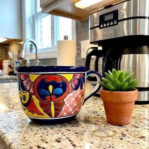 Op de afbeelding: Een grote, handbeschilderde keramische mok met een levendig bloemmotief in geel, rood, blauw en roze. Een kleine terracotta pot met een groene vetplant staat naast de mok op een gespikkelde aanrecht. Een koffiezetapparaat staat op de achtergrond.