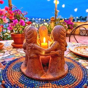 May include: A brown clay candle holder featuring a circular group of four stylized figures with arms linked, surrounding a central lit yellow candle. The sculpture has a textured, earthy finish and sits on a round, woven blue and orange mat.