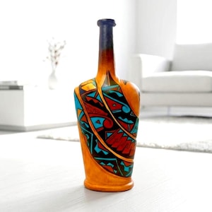 May include: A tall, decorative bottle with a unique design. The bottle features a tan base with a dark blue neck and is adorned with a colorful pattern of red, turquoise, and black geometric shapes. The bottle is standing on a white surface.