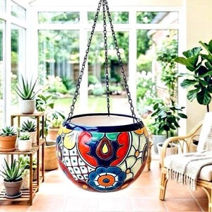 May include: A round ceramic hanging planter suspended by three black metal chains. The exterior features a vibrant, hand-painted pattern with bold red, blue, yellow, and green floral and geometric motifs against a cream-colored background.