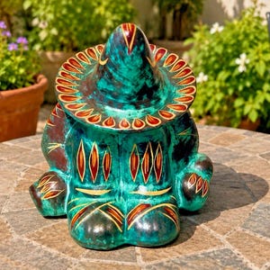 May include: A teal ceramic figurine of a seated figure wearing a wide-brimmed hat. The piece features a mottled green and blue glaze with hand-painted red and gold leaf-shaped patterns decorating the hat brim, torso, and feet of the sculpture.