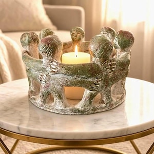 May include: A circular candle holder featuring a ring of stylized figures with arms linked around a central cream-colored pillar candle. The figures have a textured, weathered green and white finish, creating a sculptural base that encircles the light.