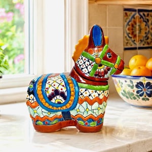 Peut inclure: Une figurine d'âne en céramique avec une finition brillante et des motifs peints à la main aux couleurs vives bleu, orange, vert et jaune. L'âne est orné d'une selle, d'un harnais et de motifs floraux et géométriques complexes sur tout le corps.