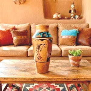 May include: A Southwestern-style living room with a tan sofa, decorative pillows, and a wooden coffee table. A tall, hand-painted vase with geometric and figurative designs is the focal point. A small potted plant sits on the table.
