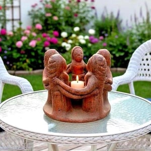 May include: A circular terracotta candle holder featuring a group of five stylized figures standing in a ring with their arms linked around a central white pillar candle. The sculpture has a textured, earthy brown finish and a solid base.