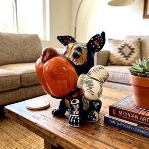May include: A ceramic dog figurine with a large orange snout and black and white accents. The dog has a colorful floral design on its face and ears. The figurine is sitting on a wooden table.