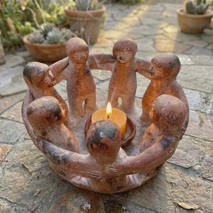 May include: A terracotta candle holder featuring six figures in a circle, holding hands. A lit candle sits in the center. The figures are a reddish-brown color. The candle holder is on a stone surface.