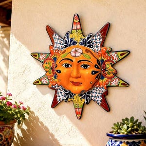 May include: A hand-painted ceramic sun wall plaque featuring a central orange face with detailed facial features. The sun rays are decorated with intricate floral patterns and geometric designs in vibrant shades of red, yellow, blue, and green.