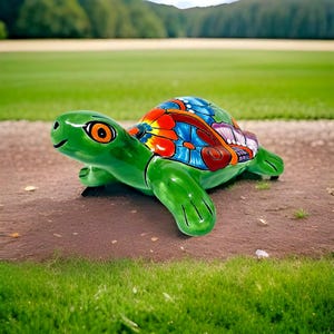 May include: A brightly coloured ceramic turtle ornament. The turtle is predominantly green with a shell decorated with red, blue, and orange floral patterns. It has a smiling face and orange eyes. The ornament is placed on a dirt path with a green grassy background.