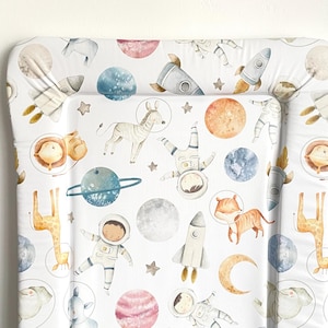 May include: A white changing mat with a space-themed print. The print features astronauts, planets, stars, rockets, and animals such as giraffes, zebras, and tigers.