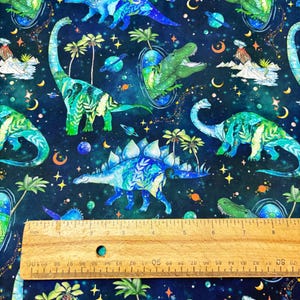 May include: A dark blue fabric with a repeating pattern of watercolor dinosaurs, planets, stars, and palm trees. The dinosaurs are in shades of blue and green. A wooden ruler is at the bottom of the image, with inch measurements.
