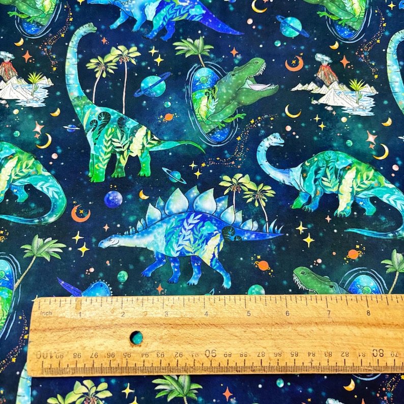 May include: A dark blue fabric with a repeating pattern of watercolor dinosaurs, planets, stars, and palm trees. The dinosaurs are in shades of blue and green. A wooden ruler is at the bottom of the image, with inch measurements.