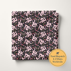 Op de afbeelding: Een gevouwen stof met een herhalend patroon van lichtroze slangen, kleine roze bloemen en gele maansikkels tegen een donkergrijze achtergrond. Het ontwerp is consistent over het gehele zichtbare oppervlak van het textiel.