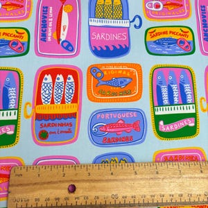 May include: A light blue fabric featuring a repeating pattern of colorful sardine and anchovy tins. The tins are in various shapes and colors, with text in multiple languages. A wooden ruler is placed across the fabric, measuring approximately 6 inches.