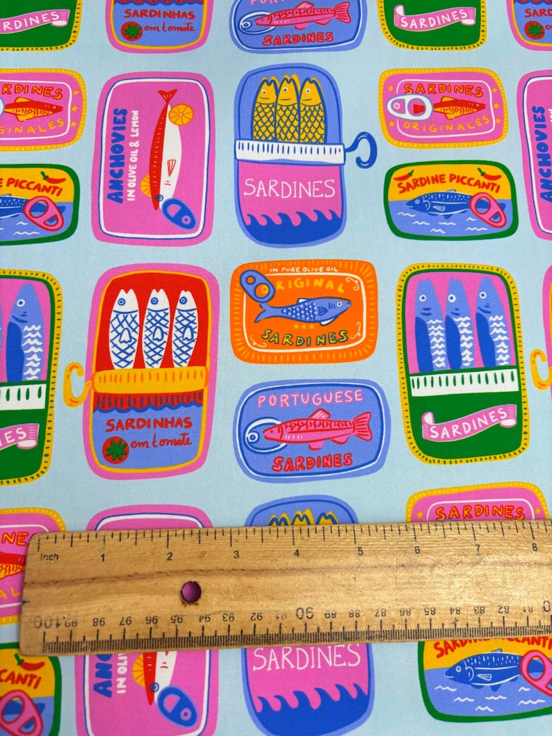 May include: A light blue fabric featuring a repeating pattern of colorful sardine and anchovy tins. The tins are in various shapes and colors, with text in multiple languages. A wooden ruler is placed across the fabric, measuring approximately 6 inches.